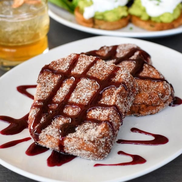 Two square churros, dusted with sugar and drizzled with red berry sauce, on a white plate.
