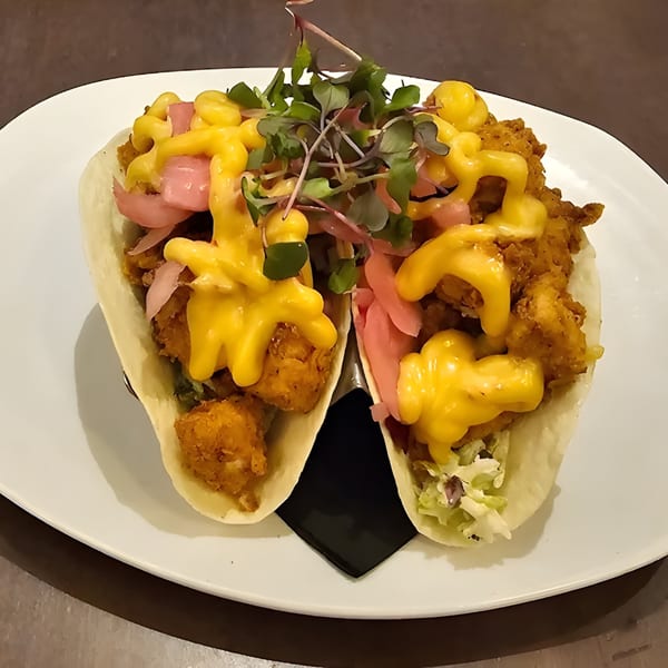 Two crispy chicken tacos with yellow sauce, pickled onions, and microgreens on a white plate.