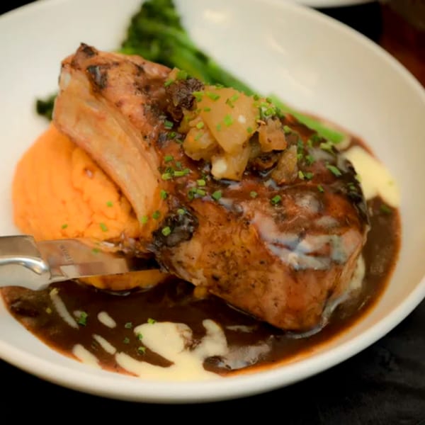 Grilled pork chop with apple chutney, sweet potato mash, gravy, and broccolini on a white plate.
