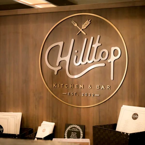 Hilltop Kitchen & Bar's circular logo with a fork and wheat stalk, on a wooden wall.