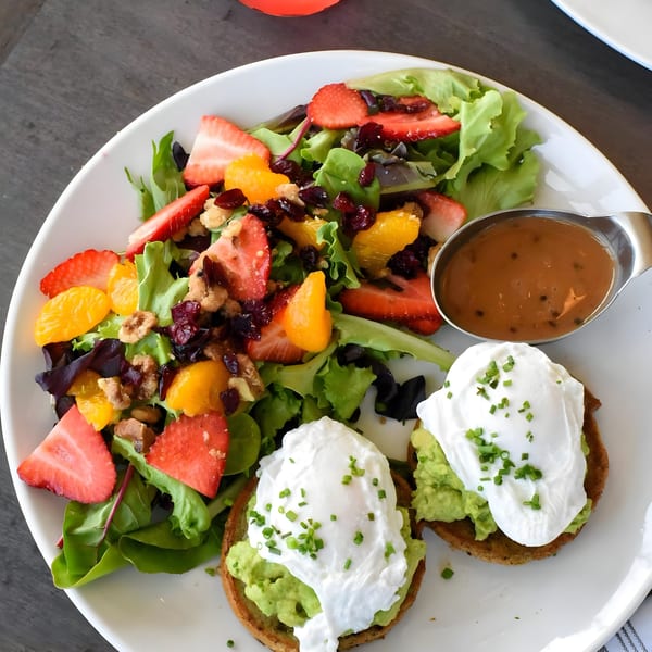 A white plate with avocado toast topped with poached eggs and chives, alongside a fruit and nut salad with dressing.